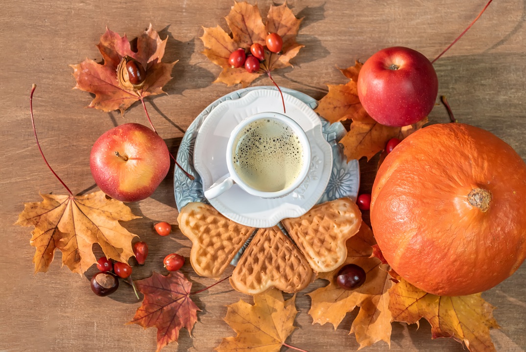 Image of wooden table with Autumn-themed items including colored leaves, apples, and a pumpkin, along with a cup of tea and heart shaped waffle cookies