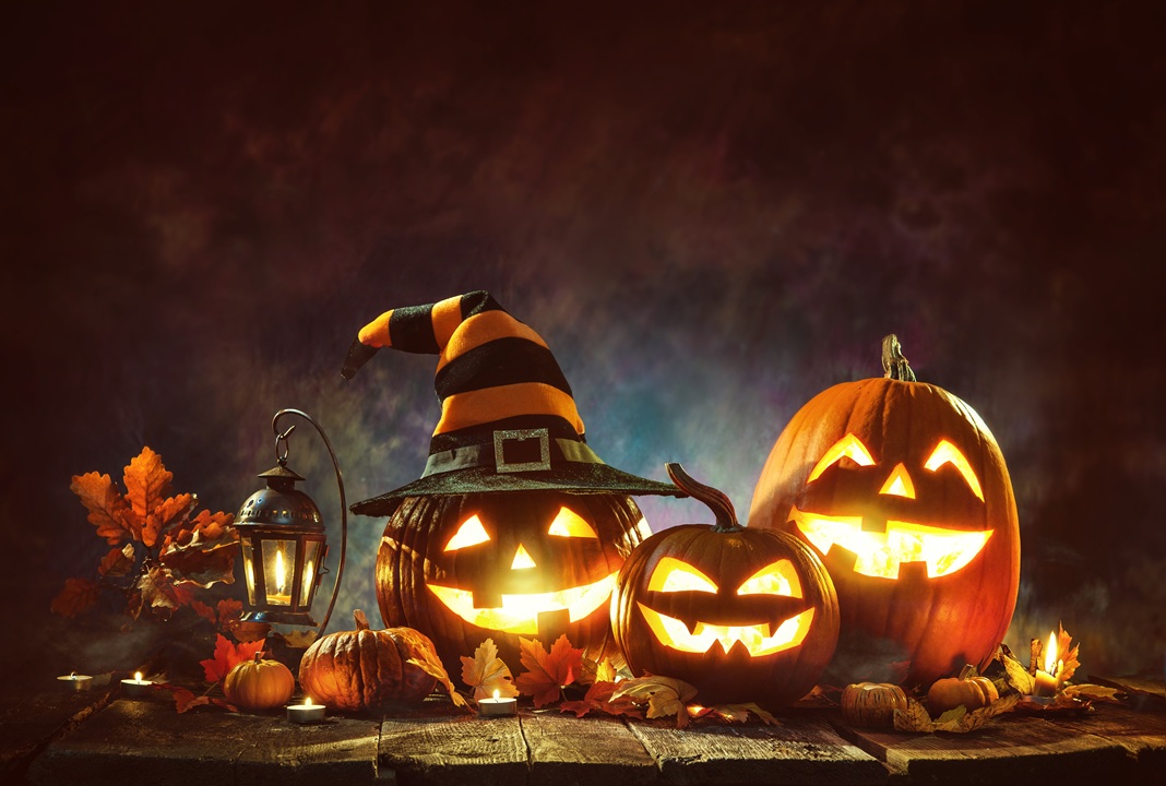 Image of three carved pumpkins, lit from inside, one wearing a hat, sitting on a table along with a lantern and small pumpkins and fall colored leaves. Dark background makes the lit candles glow.