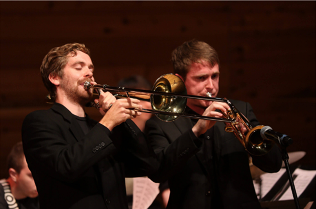 Photo of two students wearing black and performing at a concert, one playing trombone, the other playing trumpet