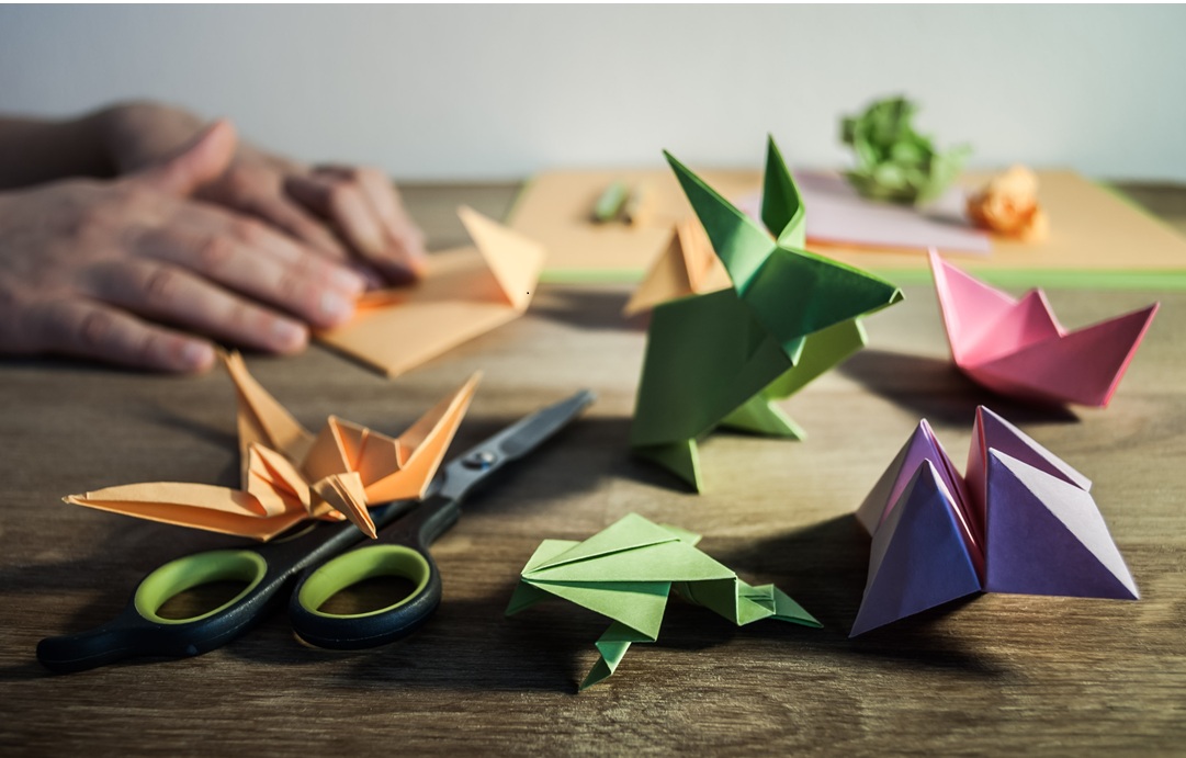 Image of hands doing origami by folding colored construction paper, scissors in background