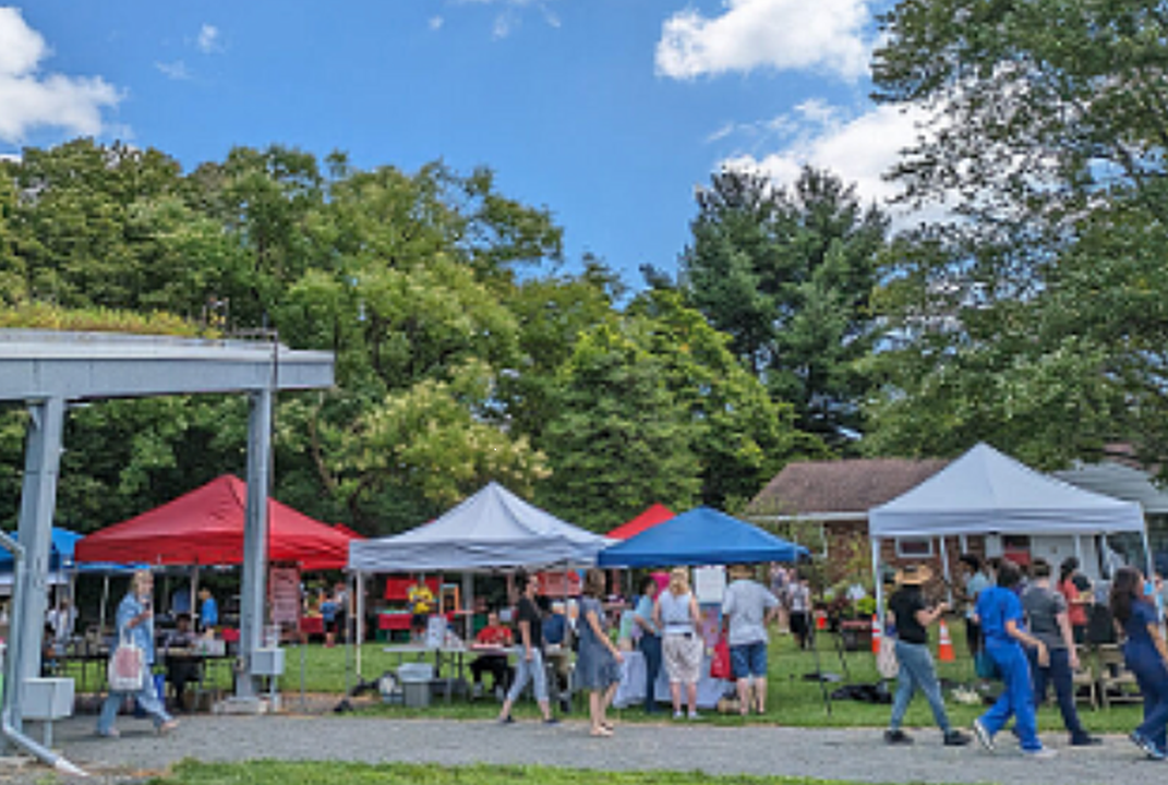 Image of people shopping at a farmers market at Rutgers Gardens. Red, white and blue canopy tents are set up over vegetable stands. Trees and blue sky in background