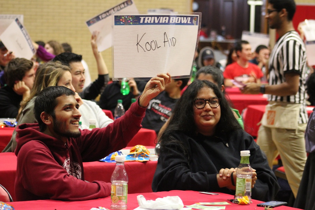 Photo from prior Trivial Bowl event, with images of people seated at tables playing trivia game. In the forefront, a male is holding up a sign with his answer to the trivia question written on it (says Kool Aid). In the background are other participants sitting at tables and a student wearing a referee shirt working the event.