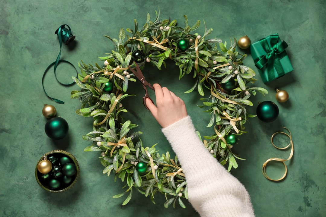 Photo of a person's arm as it works on crafting a holiday wreath. Green table in background, with a round wreath of greens on it, along with ribbons and ornaments to decorate the wreath. The person is holding scissors and wearing a white sweater.