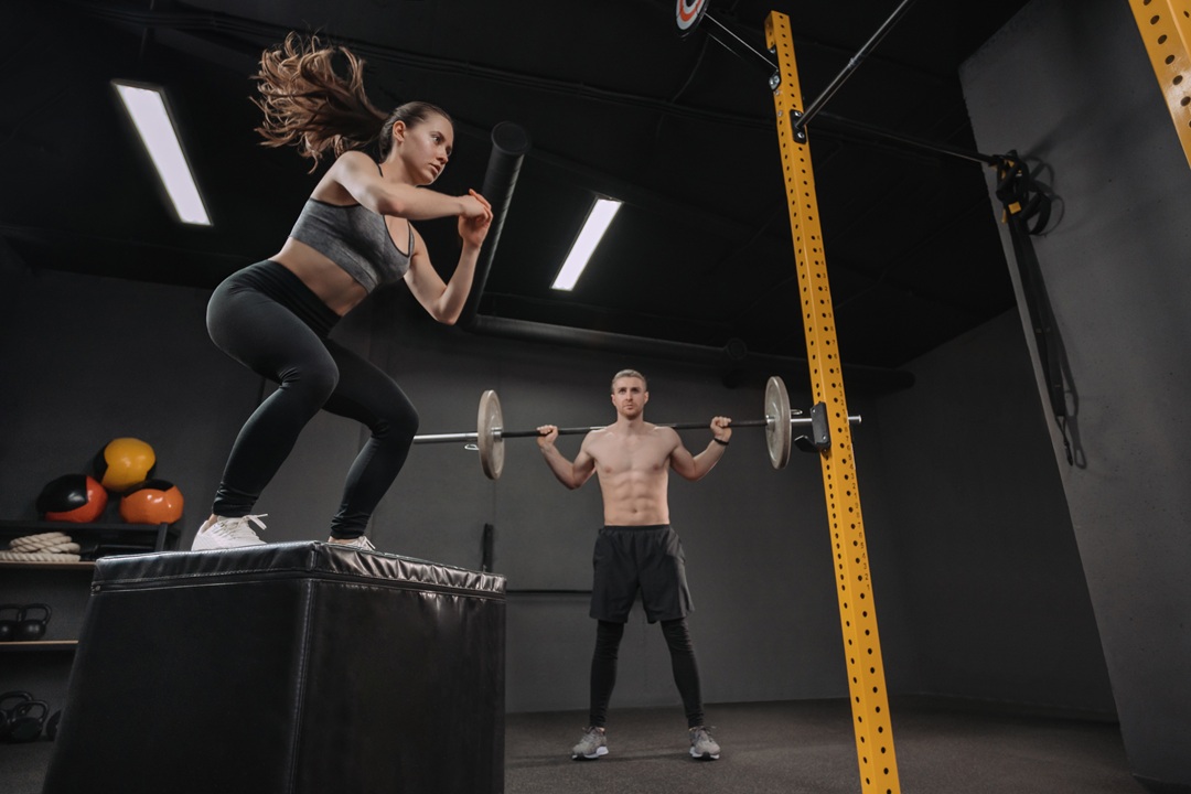 Photo of two people exercising in a gym - a woman is doing a squat on a squishy block, a man is lifting a weighted barbell