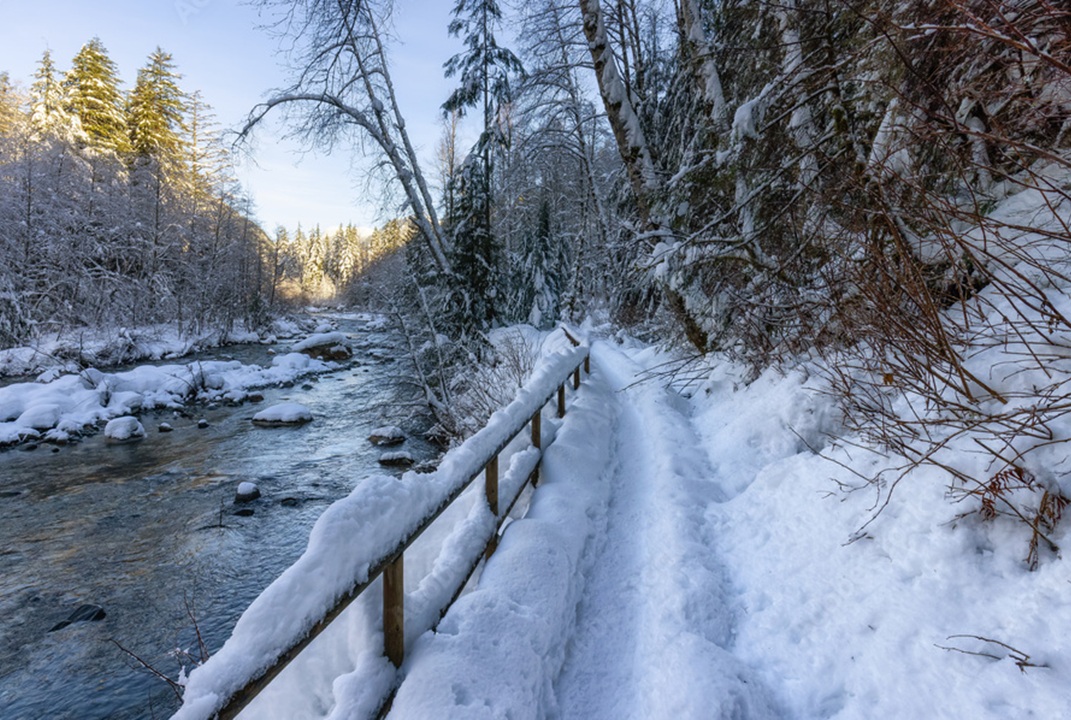 Photo of a trail, snow-covered and with a split rail fence and running water along the left side and woods on the right side