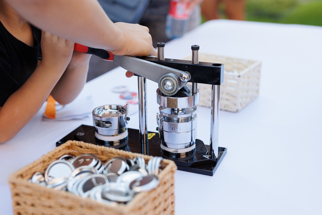 Photo of an adult assisting a child to push down on the lever of a metal button making machine. Basket of blank buttons on one side, empty basket on the other side.