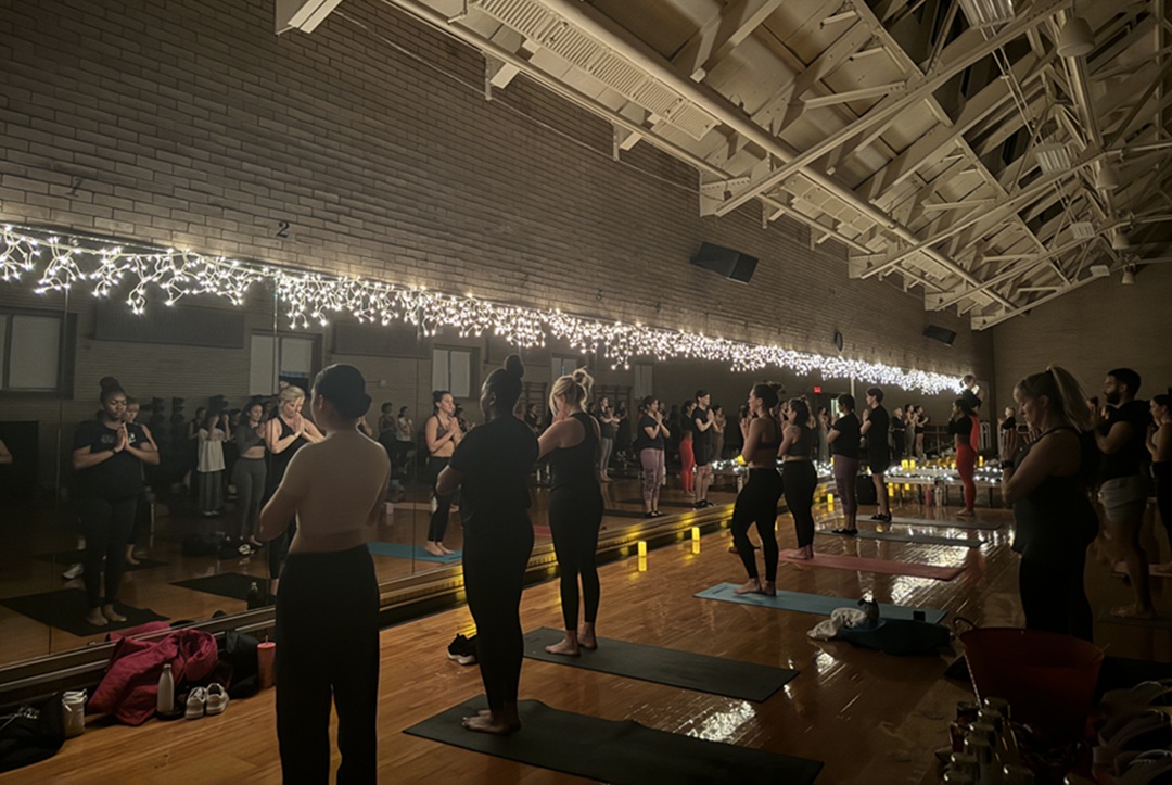 photo of yoga class with in a dimly lit gym studio with candles lit and string lights, and several students standing at front of their mats with hands in prayer at heart (mountain pose).