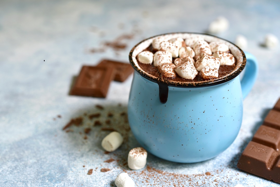 Photo of a blue mug filled with hot chocolate and mini marshmallows, on a table with chocolates and mini marshmallows and cocoa dust scattered on it.