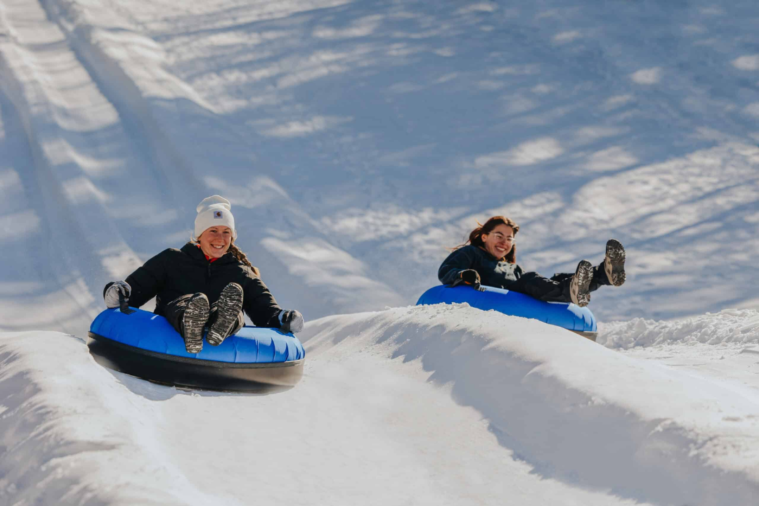 Photo of two young adults snowtubing down a mountain on a cold sunny day