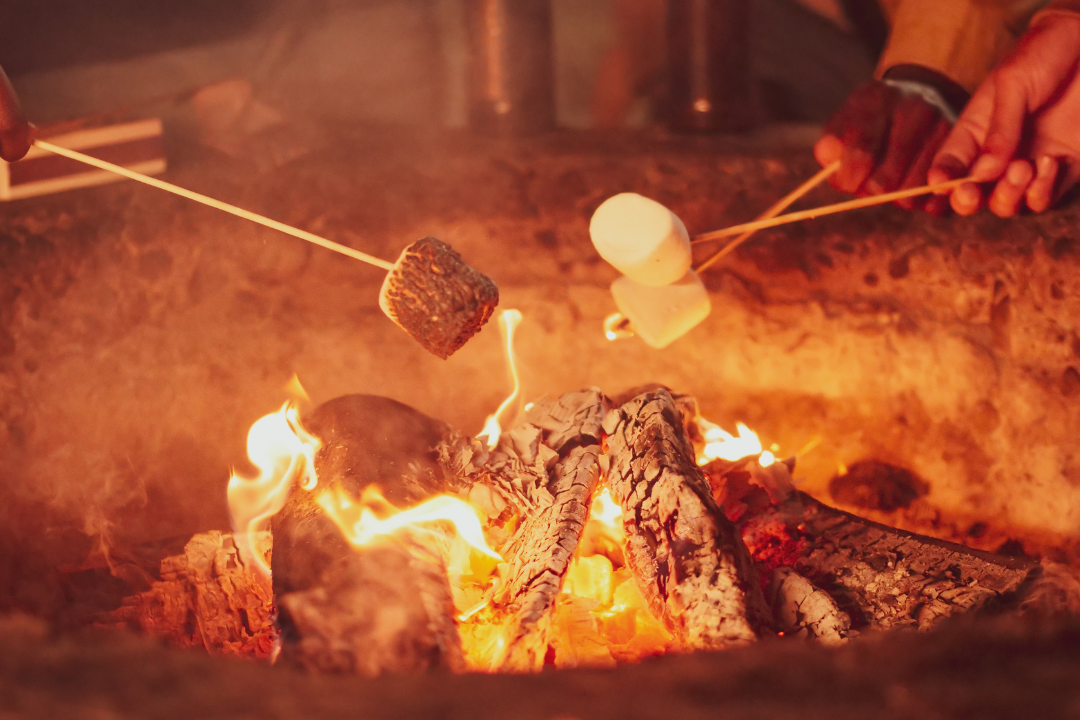 Photo of three people roasting marshmallows on a stick around a campfire, at night