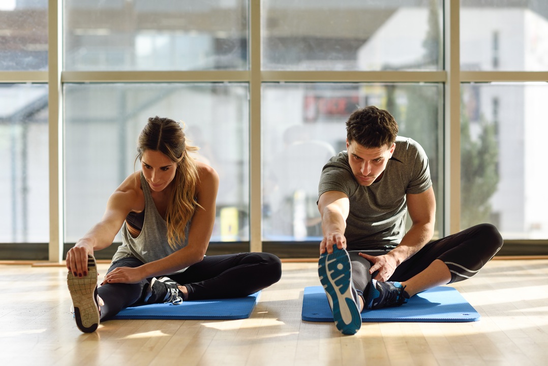 Photo of a woman and a man sitting on a mat and stretching their legs, in an exercise studio with large windows in background