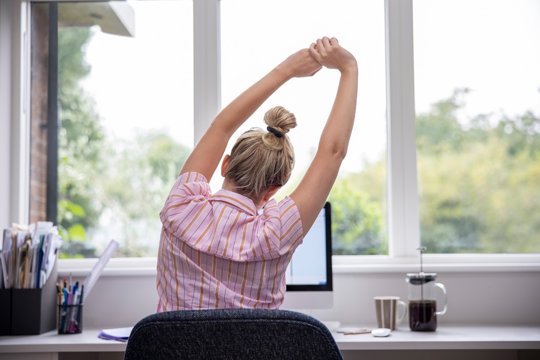 Photo of a woman sitting in a chair at her desk in front of a computer and looking out the window while she stretches her arms overhead and leans to the side