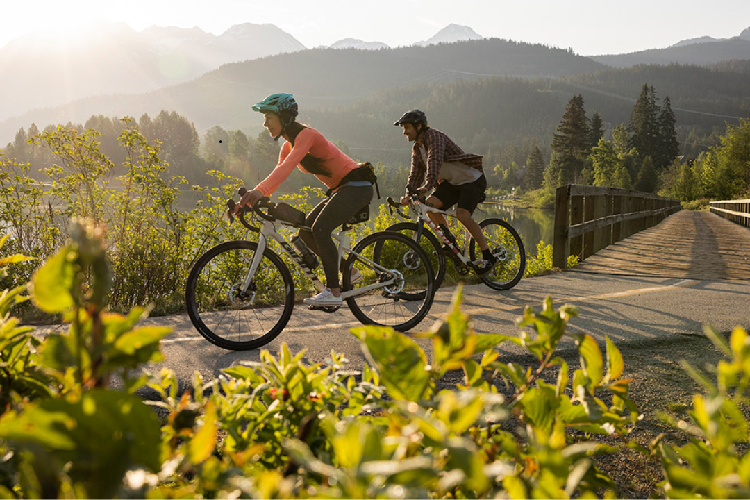 Photo of a woman and a man on bicycles, riding on a trail with flora and mountains in the background