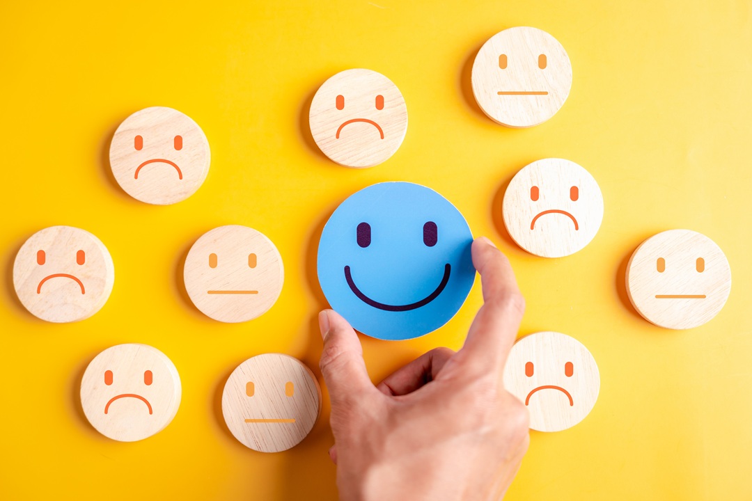 Image of round wooden discs with happy, sad and neutral faces shown on them. Yellow background, beige faces except for the smiling face with is blue and larger than the others