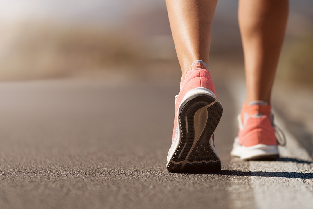 Photo of the bottom half a of a woman's legs, and feet wearing sneakers, while she walks away from the camera down the middle of a paved road.