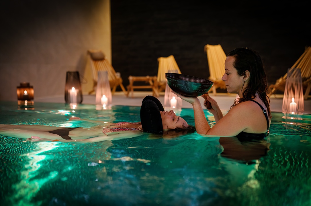 Photo of a woman floating in a pool with another woman holding a bowl over her creating a sound bath experience. The background shows darkness with candles around the edge of the pool.