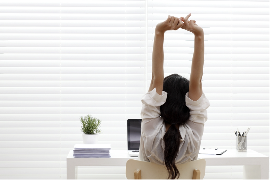 Photo of a woman sitting at a desk and stretching her arms overhead. The woman is facing away from camera, toward a window with the blinds drawn.