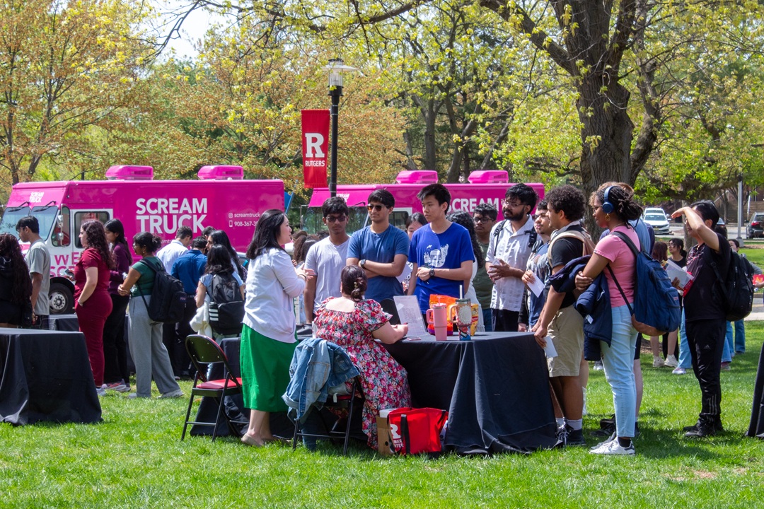 Photo of students gathered at an information table set up in a grassy area, with pink ice cream trucks in the background.