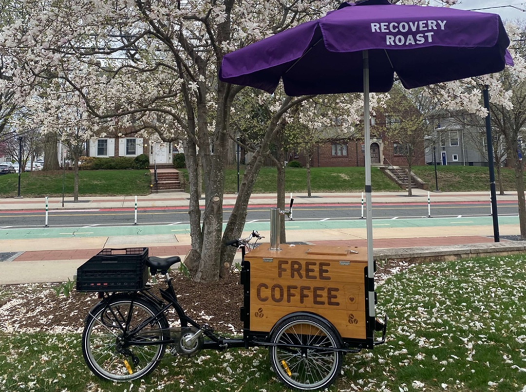 Image of the Rutgers Vicious Cycle coffee bike that is ridden around campus distributing free coffee and talking with students about addiction and recovery. Purple umbrella attached to the coffee cart reads "Recovery Roast"