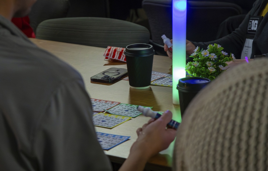 Photo of the back of a person sitting at a table playing bingo with others, stamp marker in hand, with a glow lamp and cup of coffee in the background