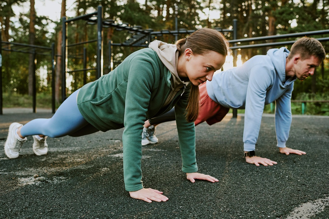 Photo of a young woman and a young man doing push-ups outdoors in a park, wearing athletic clothing