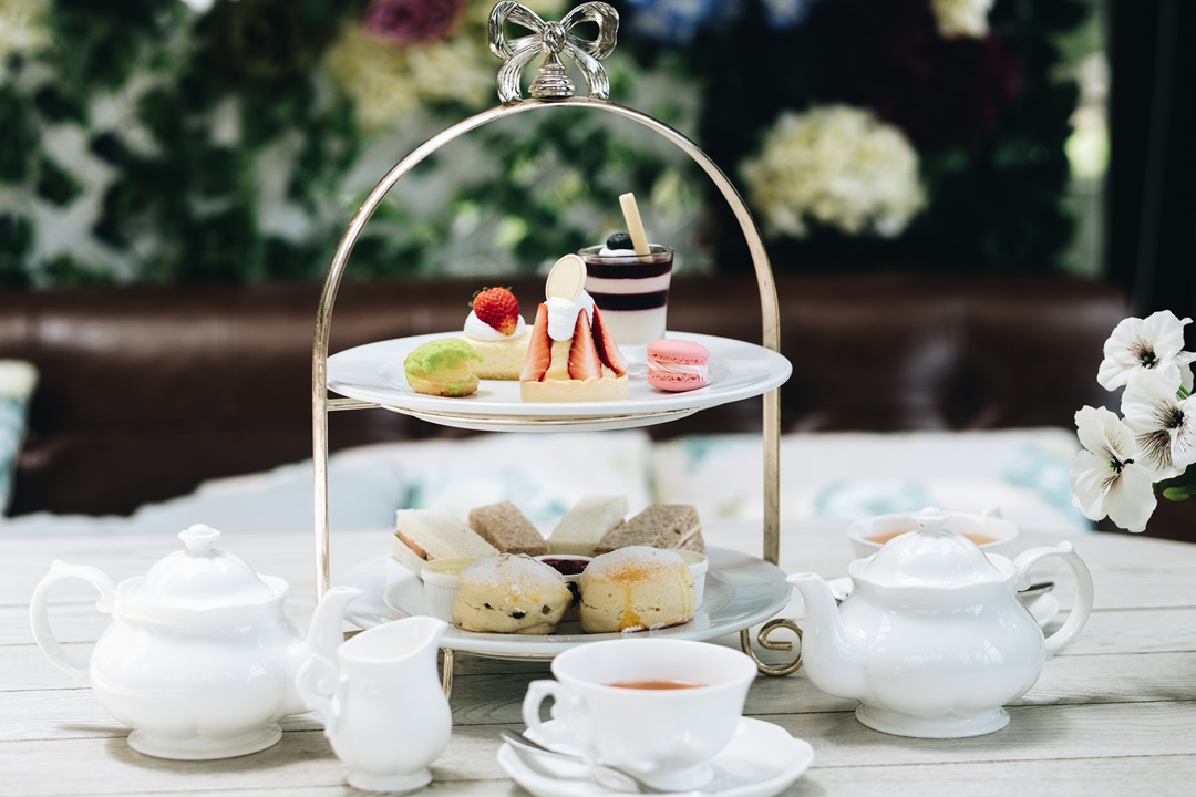 Photo of a high tea setup, on a table with flowers in the background. The setup consists of a white teapot, teacups and saucers, and a tiered rack for pastries and sandwiches.