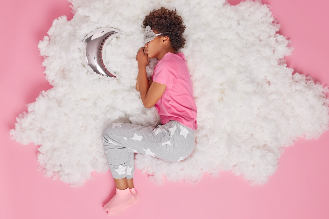 photo of a young lady laying down on her side, on a pink background with fluff around her, an eye mask on, and a silver crescent moon beside her. She is wearing a pink t shirt and grey sweatpants with stars on them.