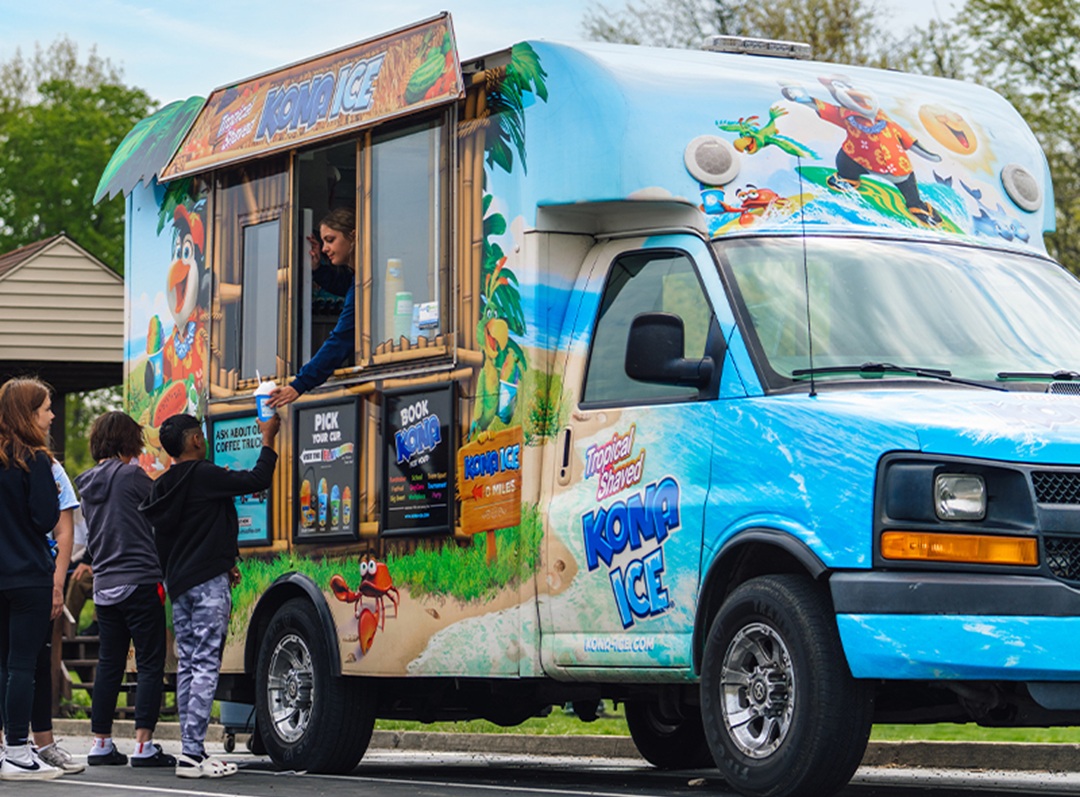 Photo of an ice cream truck serving Kona Ice to a line of people. The truck is sky blue with lots of brightly colored images painted on it, and the side of the truck renovated to sell and serve their product through a window.