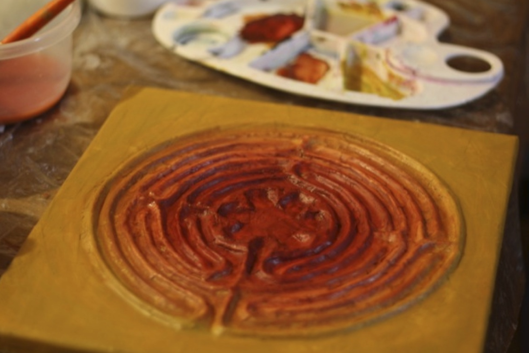 Photo of a handmade finger labyrinth, with a ceramic paint palette in the background with earth tone colors of brown and gold.