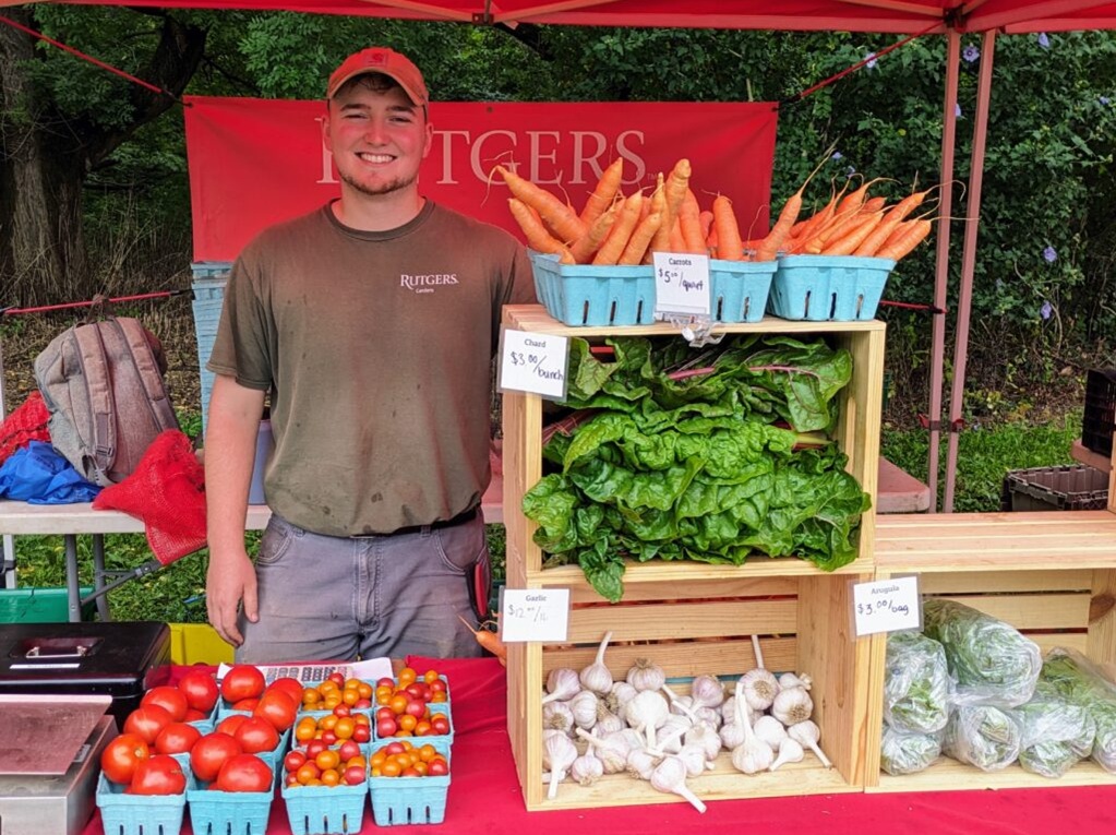 Photo of a young man standing behind a table with fresh carrots, tomatoes, garlic, and greens available for purchase.