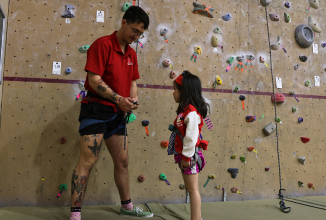 Photo of a male student employee at Rutgers Recreation speaking with a young girl about rock wall climbing. Image of rock wall is behind the two.