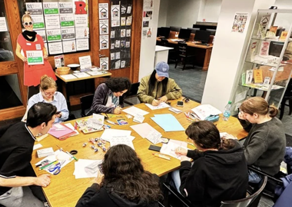 Photo of a group of students seated around a table with art supplies, colored paper and magazines spread out across the table. They are making mini magazines called zines with these materials.