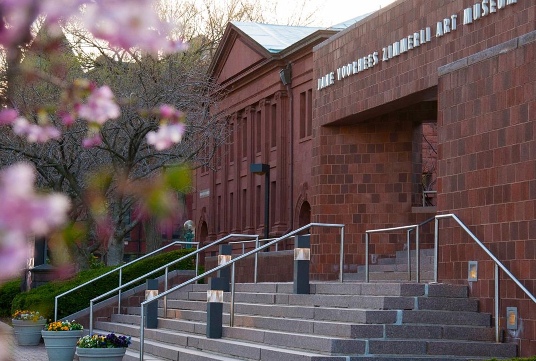 Photo of the exterior of the Zimmerli Art Museum, from the base of the steps leading into the museum. There are flower pots on the ground and a blurred image of a pink flowering tree to the left of the building.