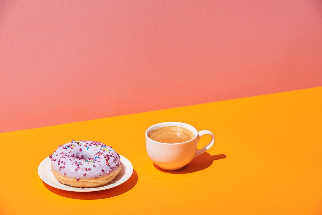 Image of a cup of coffee and a donut on a plate, with pink and orange background