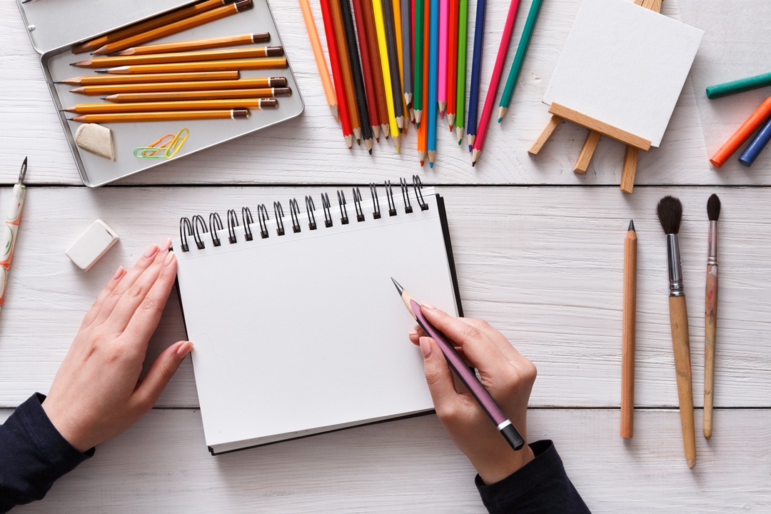 Photo of a woman's hands holding a sketch pencil in one hand, and sketching on a pad of paper, with several sketching pencils on the table around her.