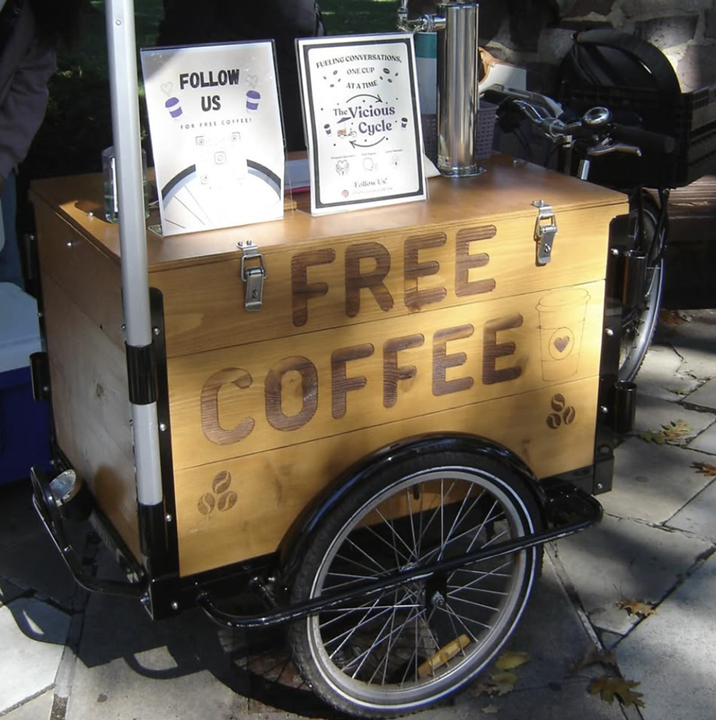 Photo of Rutgers Vicious Cycle bike, used to offer free coffee at different spots on campus, and also promote healthy habits. There is a wooden box on the front of the bike with a wheel under it, and an umbrella above it. the back half has a seat and pedals for the cyclist.