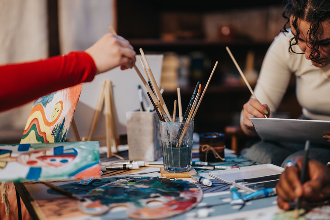 Paint brushes, paint, and art supplies on a table with two people's hands holding the brushes
