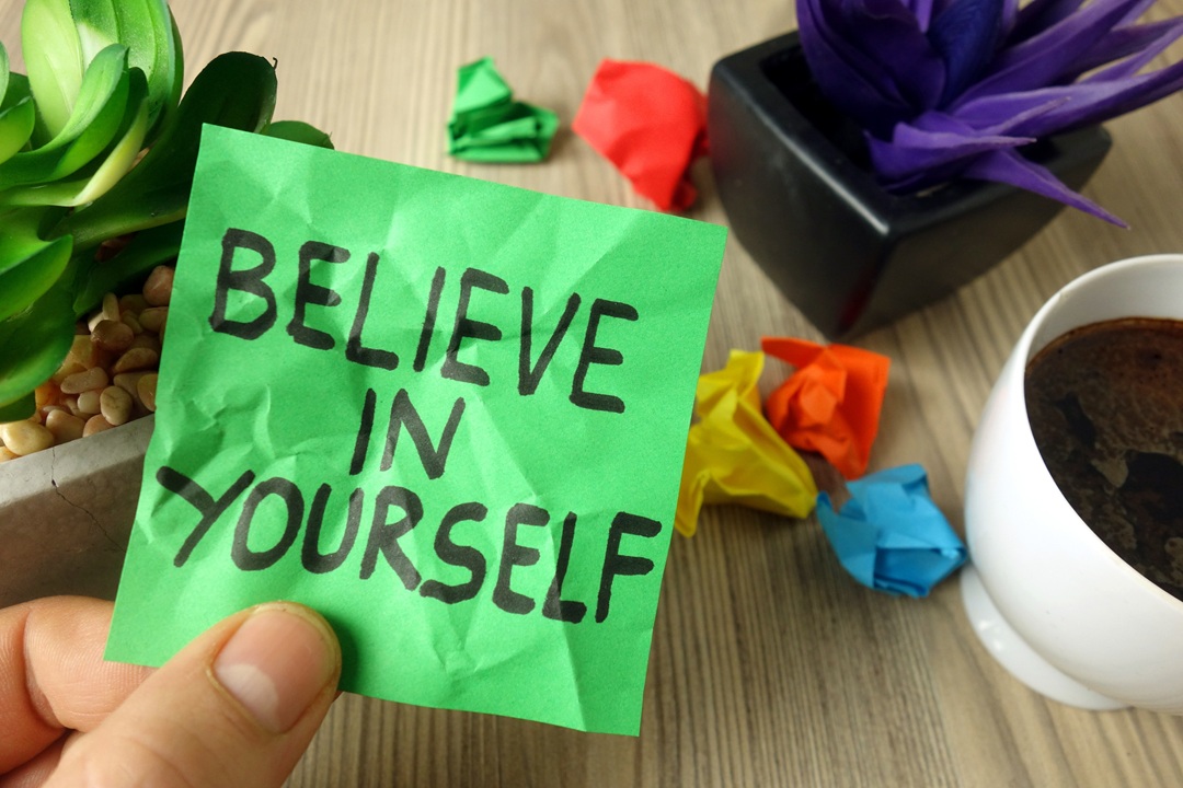 Photo of a person holding a small green piece of paper with the words Believe in Yourself written on it in black lettering.