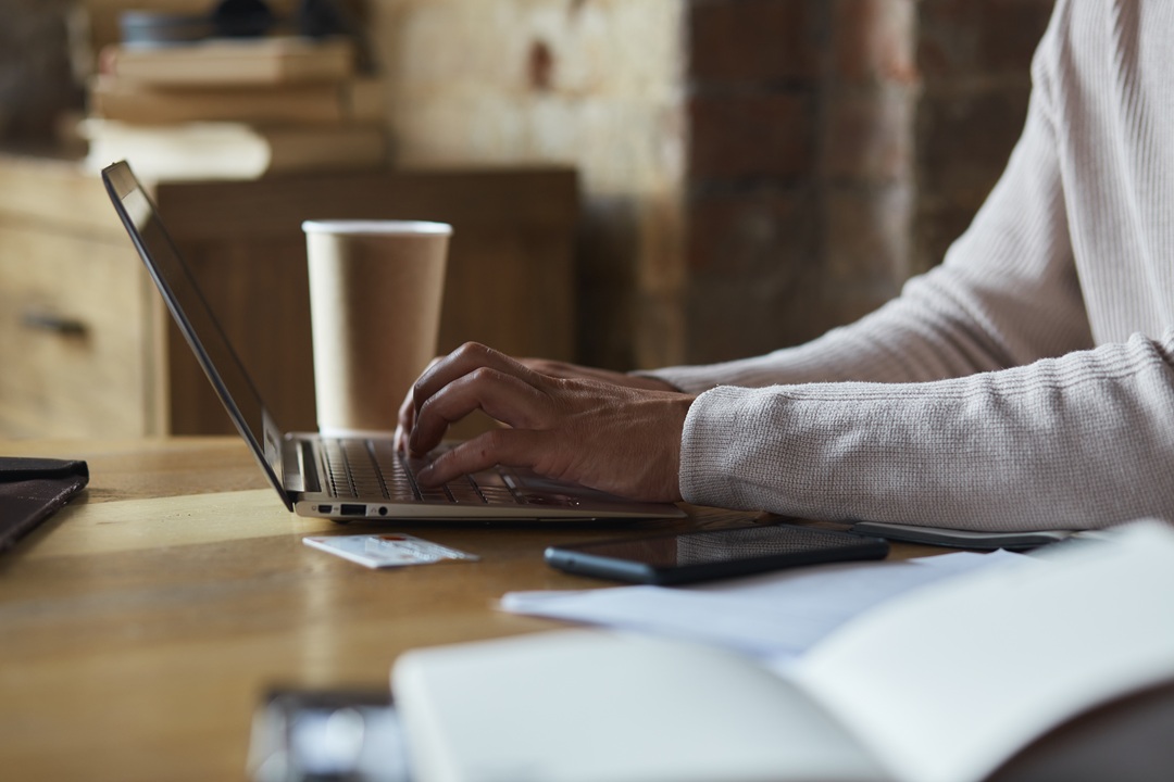 Photo of a man's hands typing on a laptop, with a cup of coffee and open books and notebooks on the table.