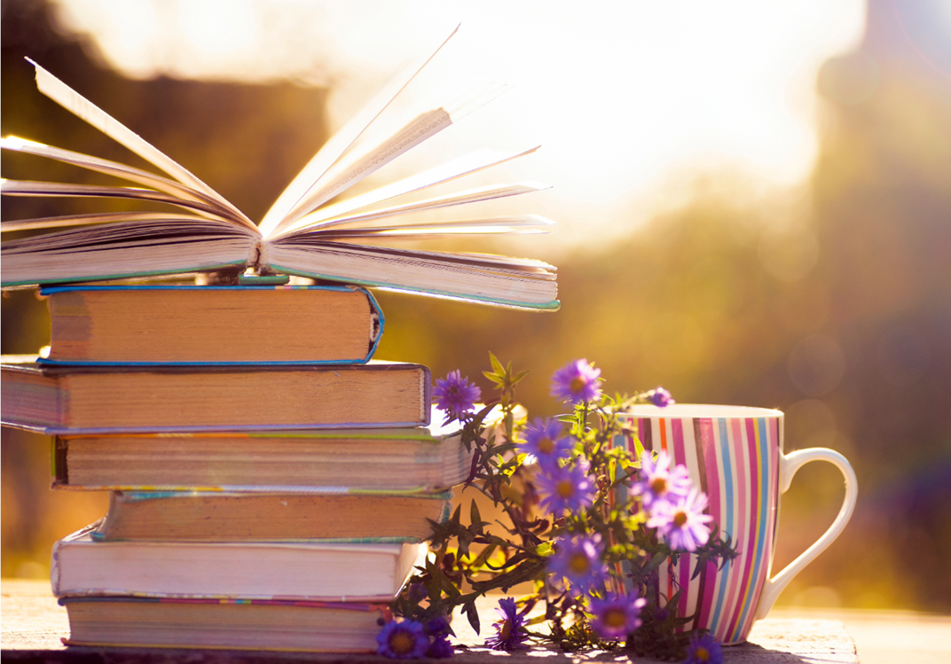 Photo of a stack of books taken outdoors, with one book fanned open on top of the stack, and a coffee cup and flower next to the stack.